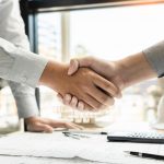 Two people in business attire shake hands over a desk with documents, a calculator, glasses, and a white construction helmet, suggesting an agreement between custom welding and fabrication experts or process vessel manufacturing professionals.