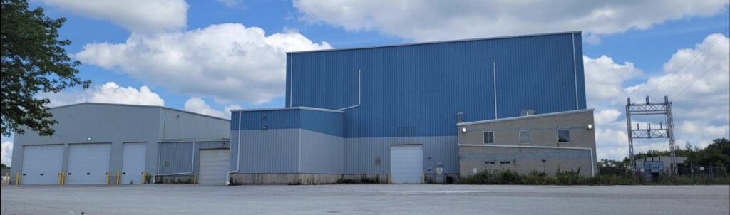 A large blue industrial warehouse with three connected buildings, ideal for custom metal fabrication, is surrounded by a wide, empty paved lot under a partly cloudy sky. Some wild grass and plants border the structure.