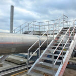 Large industrial metal pipe runs parallel to a set of metal stairs and walkway platforms, with safety railings.