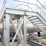 A metal platform with stairs and railings stands on concrete blocks. Large industrial pipes run underneath, and parts of the structure are supported by cross-braced beams.