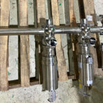 Two stainless steel industrial valves with cylindrical actuators are mounted on a horizontal pipe, placed on a wooden pallet on a concrete floor. The equipment appears new and unused.