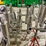 A cluster of stainless steel industrial valves and pipes is set on a workshop floor, supported by red stands. Large green cylindrical tanks and other equipment are visible in the background.