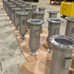 Rows of large, cylindrical metal industrial components with flanges are arranged neatly on a factory floor, with shelves, machinery, and equipment visible in the background.