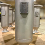 A large, cylindrical metal industrial filter vessel with a flanged base and a label plate, standing upright on a cardboard surface. Several similar vessels are visible in the background.