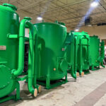 Large green industrial tanks with connected pipes standing in a row inside a spacious, well-lit facility with a high ceiling and exposed beams.