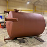 A large, cylindrical, rust-colored industrial tank with metal brackets on its side sits on wooden planks in a spacious warehouse with a high ceiling and bright lighting.