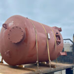A large, rust-colored metal tank is secured with straps on the back of a flatbed truck. The sky is cloudy, and industrial buildings are blurred in the background.