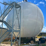 A large, shiny metal spherical tank stands outdoors, next to a tall metal staircase structure. Blue sky with clouds is visible, and construction materials are scattered around the base of the tank.